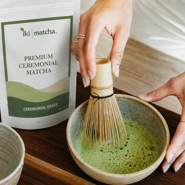 A person's hands are shown whisking green matcha tea in a ceramic bowl using a bamboo and rubber chashaku whisk. A white bag of 'iki matcha' Premium Ceremonial Matcha is visible in the background on a dark wooden table, alongside other minimalist ceramic tea accessories.