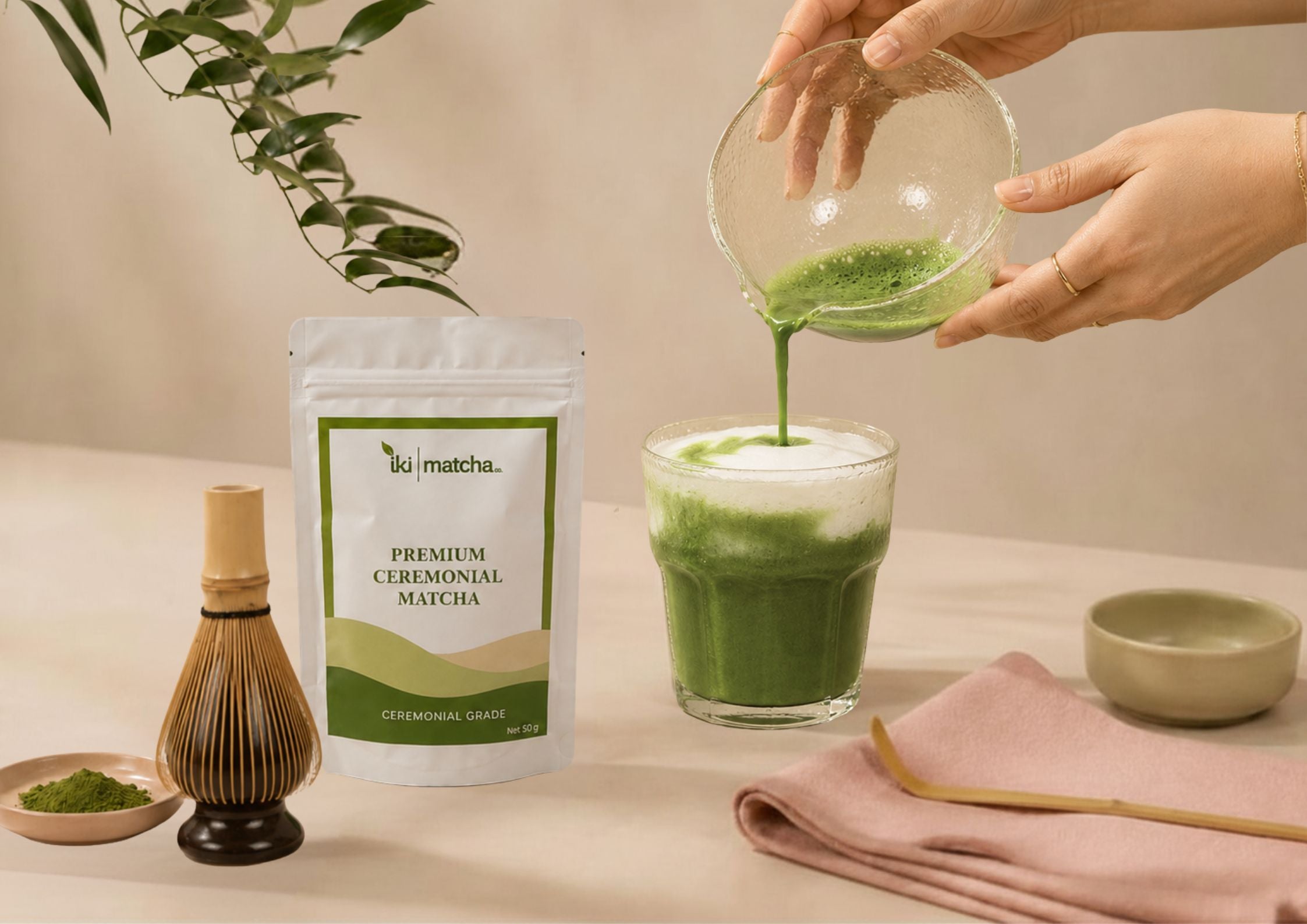 Person pouring green liquid into a glass mug with a whisk and towel on a beige background