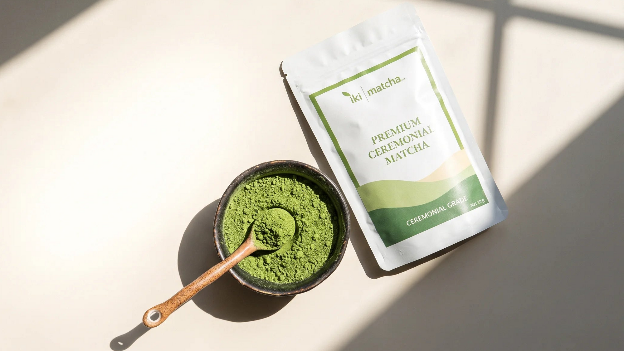 A stand-up pouch of green premium green tea powder sits beside a small bowl filled with bright green tea powder and a wooden spoon, arranged on a light surface with strong diagonal shadows.