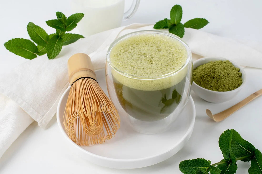 Matcha green tea with a whisk, bowl of matcha powder, and mint leaves on a white background