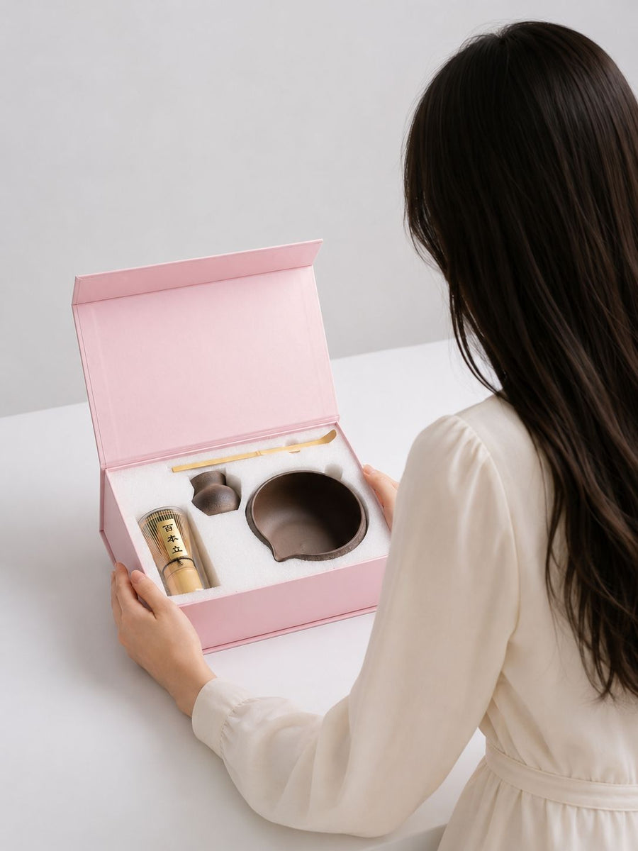 Person opening a pink gift box containing tea-making items on a white surface.