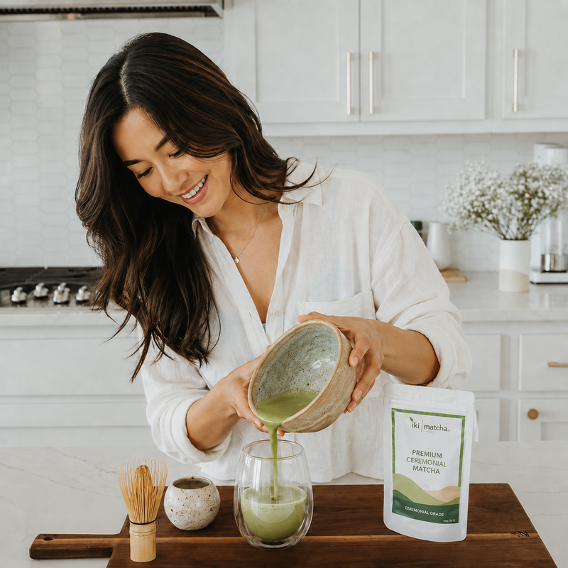 A woman in a white shirt pours green tea from a ceramic bowl into a glass on a wooden cutting board, with a pouch of matcha tea and a bamboo whisk nearby in a bright kitchen setting.